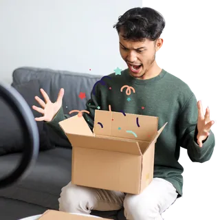 A young man in a sweater acts surprised as he opens a plain cardboard box in front of a ring light