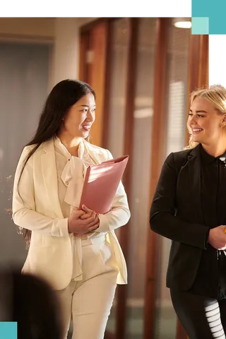 Two women in business suits smile and chat as they walk down a hallway